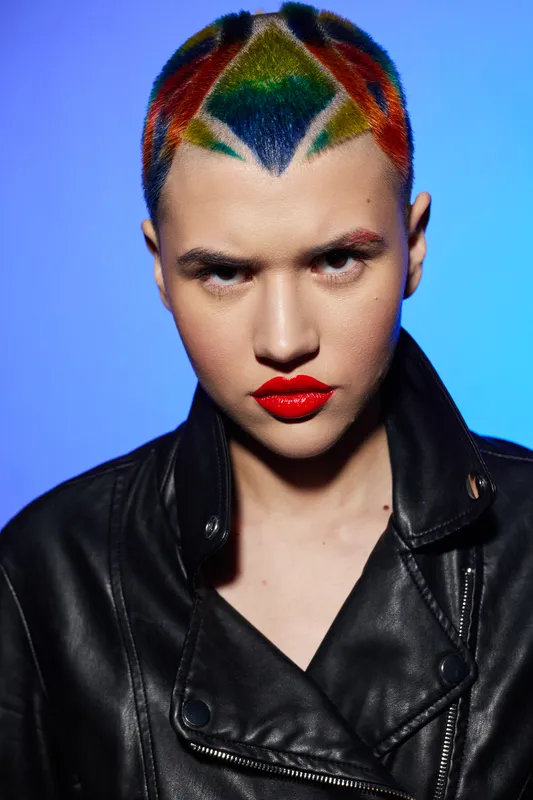 Young woman with colorful short hair and red lipstick wearing black leather jacket, staring intensely against a vivid blue background, bold style and edgy fashion.