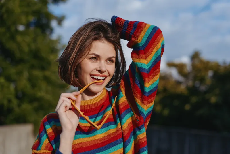 Woman smiling in a rainbow sweater holding sunglasses, casual outfit and short hair young adult outdoors, leaning back with hand in hair, lifestyle portrait, sunny mood.