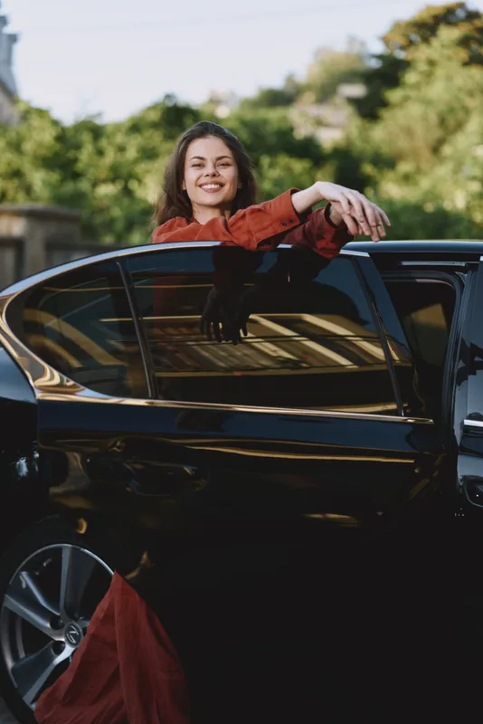 A smiling woman leans on a sleek car window in a casual red dress, capturing a candid moment of confident everyday lifestyle