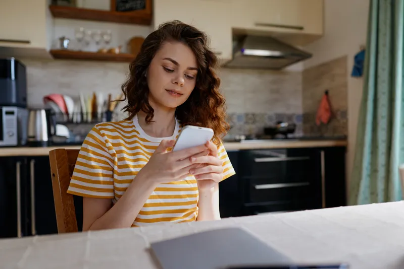 Woman at home sits in a bright kitchen, reading on her smartphone, relaxed mood, casual striped tee, cozy domestic scene, everyday life, soft natural light.