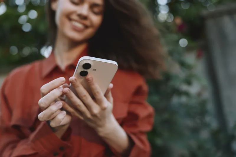 Cheerful woman outdoors, smiling with phone in hand, casual lifestyle moment, sunlit park backdrop, natural colors, relaxed pose, gentle breeze, candid everyday connectivity