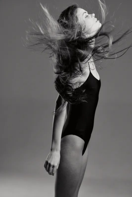 Black and white portrait of a young woman with flowing hair in a black bodysuit, expressing motion and grace with closed eyes against a gray background.