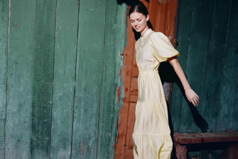 Woman in a yellow dress walks along a weathered wooden wall outdoors, posing gracefully with a subtle smile in a candid fashion moment showing rustic charm and calm elegance