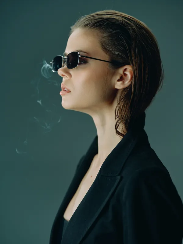 Profile of a stylish woman wearing sunglasses smoking with a green background in a moody setting.
