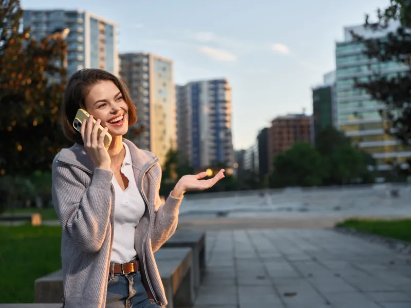 A cheerful professional woman is seen talking on a smartphone in an urban outdoor setting. She smiles warmly and appears engaged, creating a positive and dynamic business vibe for a modern workplace.