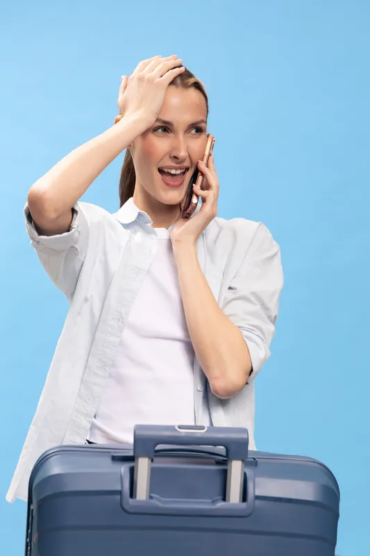 Excited young woman talks on smartphone standing behind blue suitcase on blue background. Concept of travel, vacation, surprise, and communication with casual outfit and happy expression.