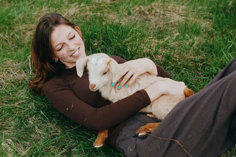 Relaxed woman lying on green grass outdoors, smiling softly as she cuddles a curious goat kid, illustrating warm companionship, rustic lifestyle, and a serene countryside moment