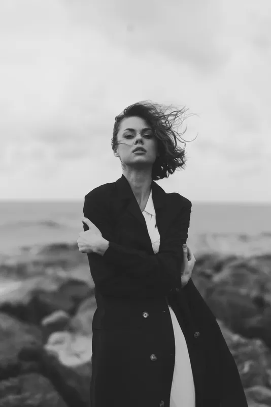 Fashion model on a windy rocky shore in a long black coat, dramatic monochrome scene, strong pose and sea breeze, editorial style fashion at the coast