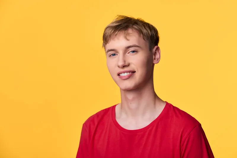 Young man in a red shirt smiles against a bright yellow backdrop, casual studio shot conveying upbeat mood, simple styling, clean composition, vibrant color contrast, energetic and approachable vibe