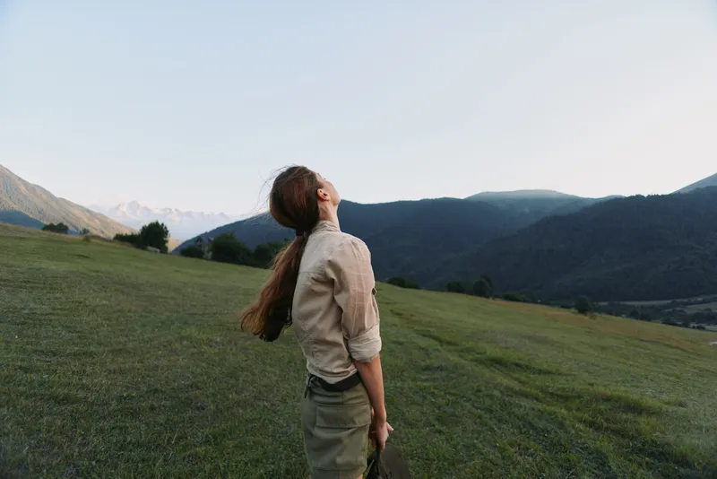 Young woman with long hair stands on a green hillside looking up at the sky surrounded by mountains during a calm outdoor moment.