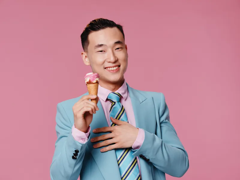 Радостный мужчина азиатской внешности держит мороженое, на нём синий костюм, на заднем плане розовый фон