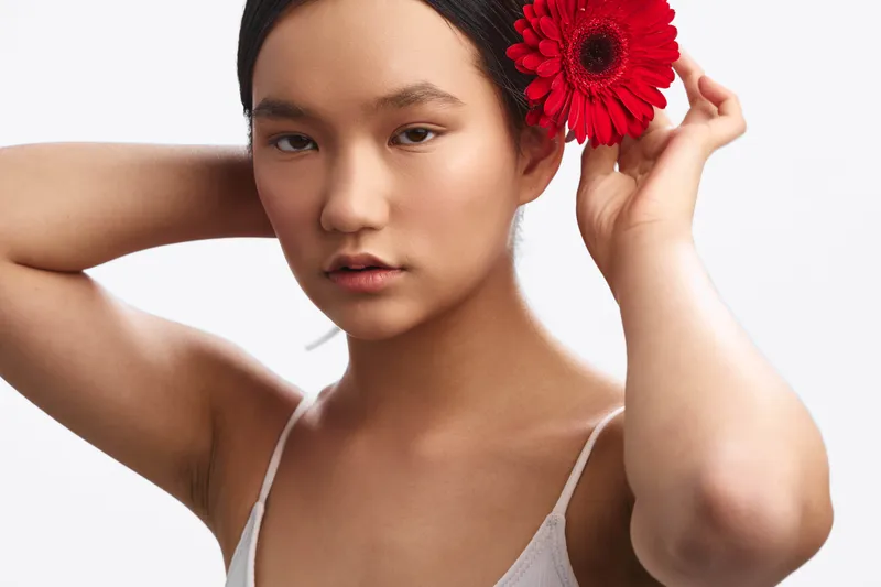 Asian woman adjusts a bright red flower behind her ear in a minimal studio portrait, conveying calm elegance, natural beauty, and a poised, contemplative mood.