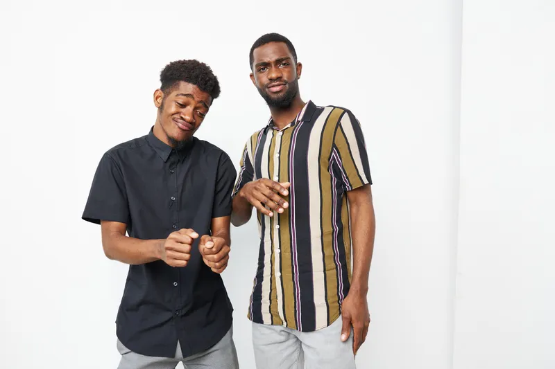 Two African American men posing confidently together, showcasing stylish outfits and a playful mood. Their expressions radiate joy and camaraderie, creating a dynamic composition.