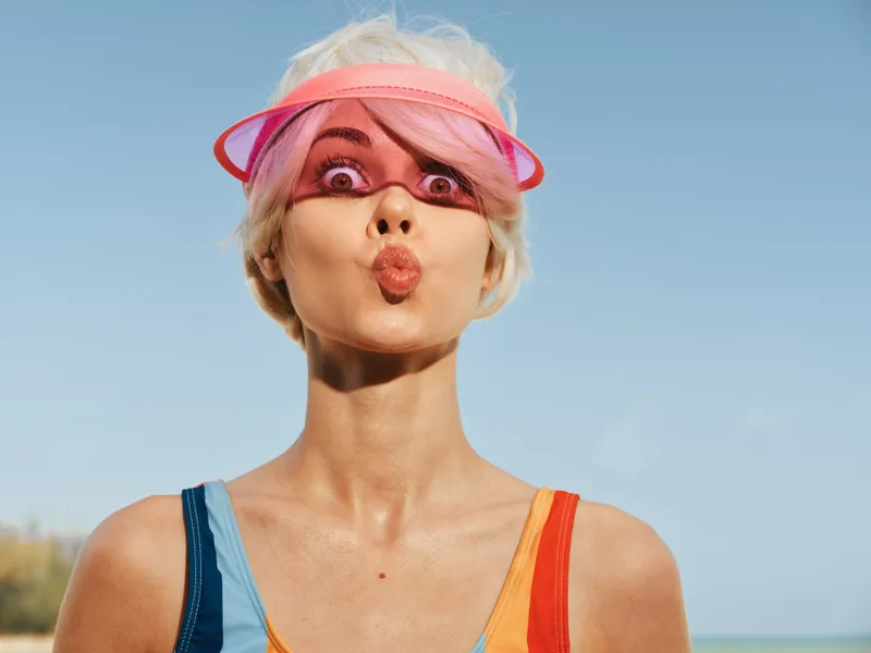 Portrait of a playful person with a pink visor, wide eyes, and puckered lips under a bright sky. Bold colors and confident pose convey energy, fun, and carefree attitude.