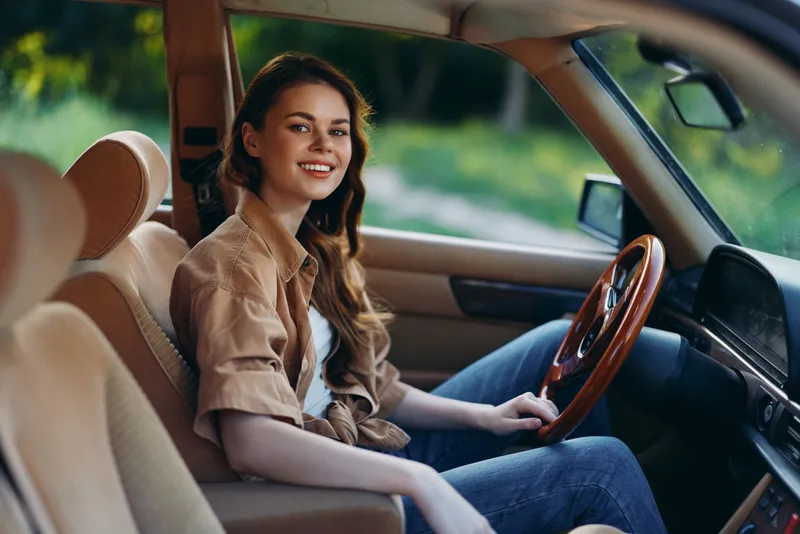 Cheerful young woman sitting in a vintage car, enjoying a summer drive in nature, wearing a casual brown shirt and smiling warmly.