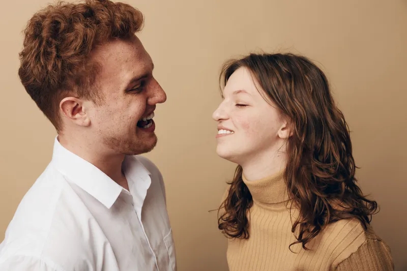 Smiling young man and woman facing each other, laughing and enjoying a happy moment together. Casual clothing and warm beige background create a friendly, joyful atmosphere.