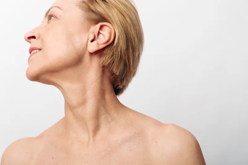 Portrait of a mature woman with short hair turning her head to the side, showing neck and shoulder against a plain white background. Concept of aging, beauty, and natural skin.