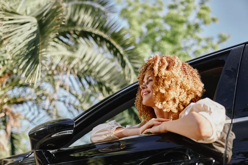 smiling woman with curly hair enjoying sunny day leaning out of car window with palm trees and blue sky background, casual and relaxed mood