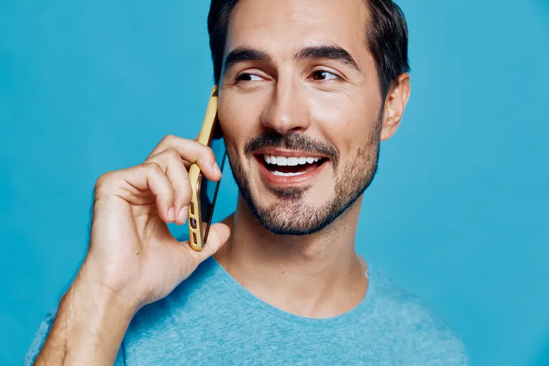 Happy smiling young man talking on mobile phone, wearing light blue t-shirt, against a solid blue background. Cheerful lifestyle concept.
