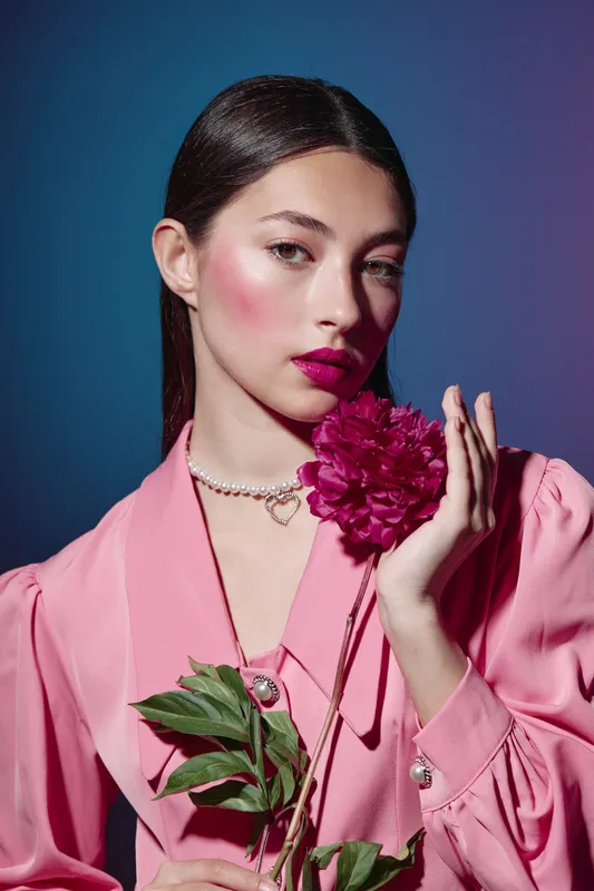 Young woman in pink oversized blouse holds deep magenta flower near her face with vibrant makeup, including glossy lips and dramatic blush. Studio beauty fashion portrait.