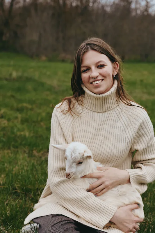 Smiling woman outdoors with a gentle goat kid in a green field, capturing a warm lifestyle moment, cozy sweater, and connection with nature, light breeze and peaceful afternoon.
