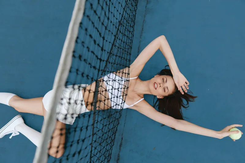 Athletic young woman in white sportswear joyfully stretching after a tennis match on a blue court, capturing the essence of sports and fitness