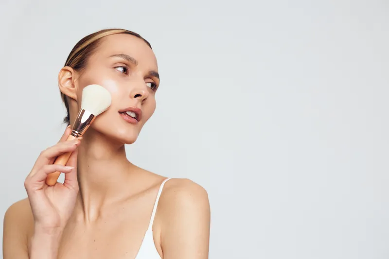 Young woman applying makeup with brush on clean skin, natural light, minimalistic background, beauty routine, focused expression, soft skin texture, casual style