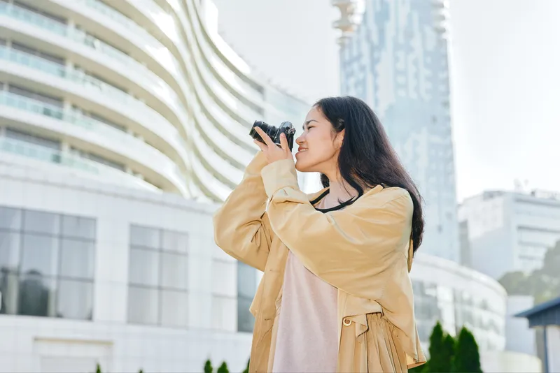 Camera, photography, woman, outdoors, urban, city lifestyle capture of a cheerful figure with a camera in hand, wearing a tan coat, bright morning light by modern buildings