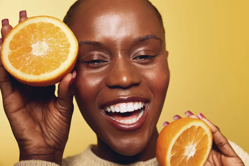 Happy smiling black African woman with dark skin holds two halves of fresh orange close to face on yellow background. Concept of healthy lifestyle, vitamin C, skincare, and natural beauty.