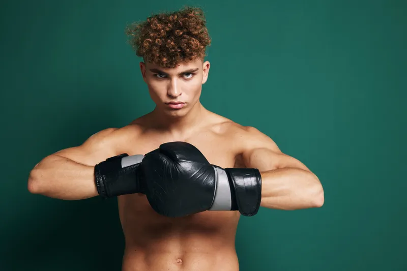 Young muscular man with boxing gloves in fighting stance training for fitness and combat sport on green background. Athlete focused on strength and discipline.