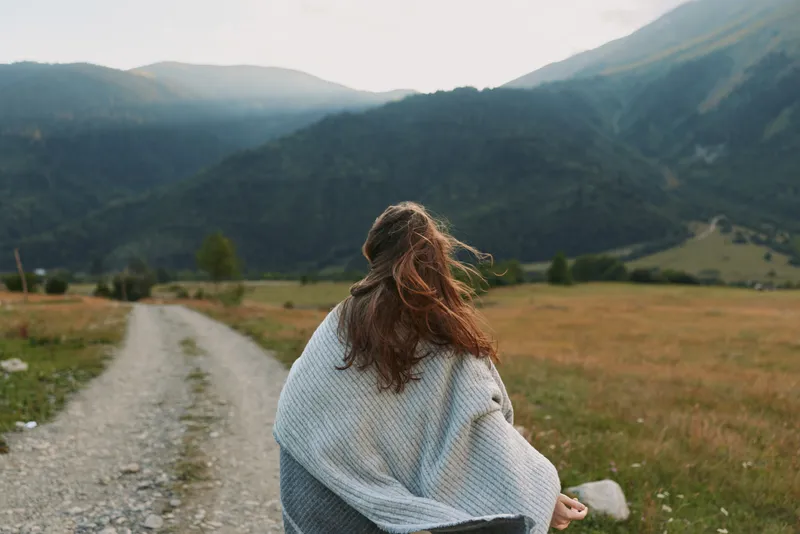 Woman from behind wearing a light shawl walks along a gravel path in a mountain valley, capturing a calm countryside lifestyle moment outdoors with wind in hair