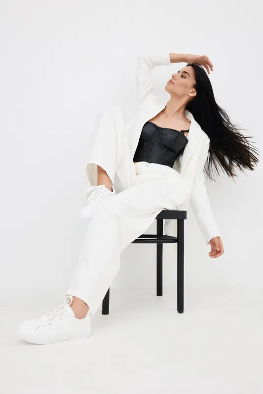 young woman sitting on black chair in stylish white pantsuit and black top with flowing hair, minimalistic studio background, modern fashion concept.