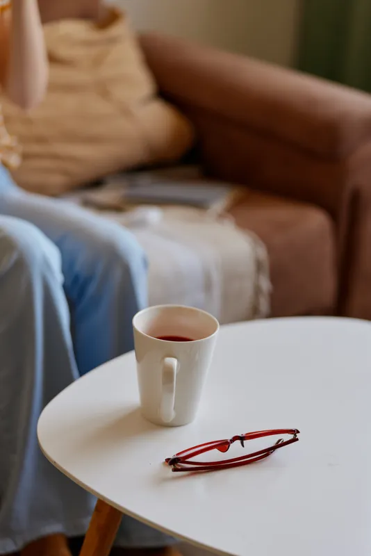 Cozy home living scene with a white coffee mug resting on a round table, reading glasses beside it, a soft blanket and sofa in the background, conveying a warm lifestyle moment in a calm room.