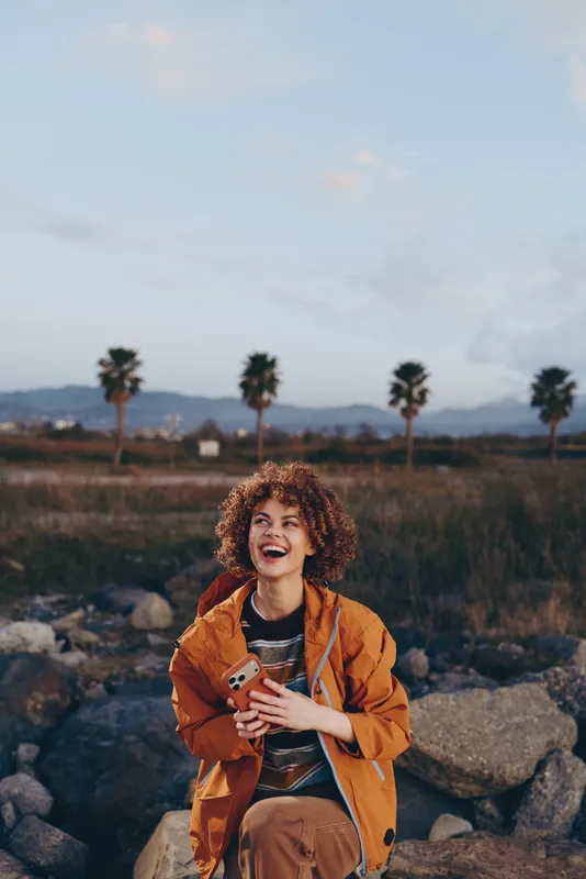 Woman lifestyle smiling curly jacket outdoors travel nature sunset palm scene, woman in orange jacket stands on rocks, palms and mountains behind, soft evening light, happy smile