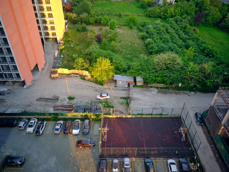Aerial view of parking with cars next to a basketball court and apartment building, green lot and construction area by a road at sunset with long shadows and warm sunlight.