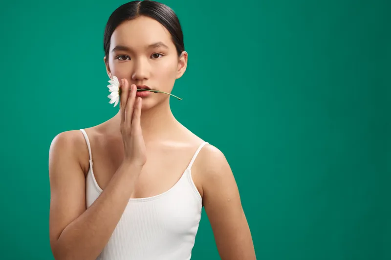 Asian woman holds a white flower near her lips in a simple studio portrait on a green background. The calm, natural mood and minimalist composition create a clean, contemporary aesthetic.