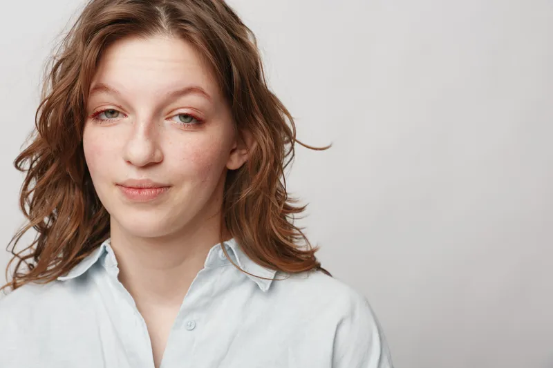 Young woman with wavy brown hair and light skin wears a white shirt, looking at camera with a subtle smile on a plain light background. Concept of natural beauty and casual portrait.