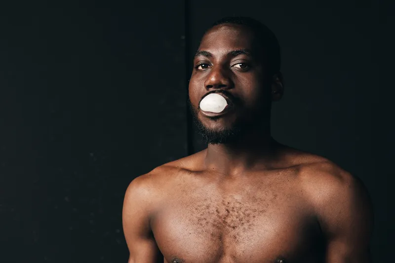 Black man with dark skin and beard in creative conceptual style holding white object in mouth against dark background. Portrait of African ethnicity with bare chest and intense gaze in studio
