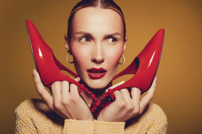 Young woman with red high heels holds shoes near face, wearing yellow sweater and hoop earrings. Studio portrait on brown background with fashion and style concept.