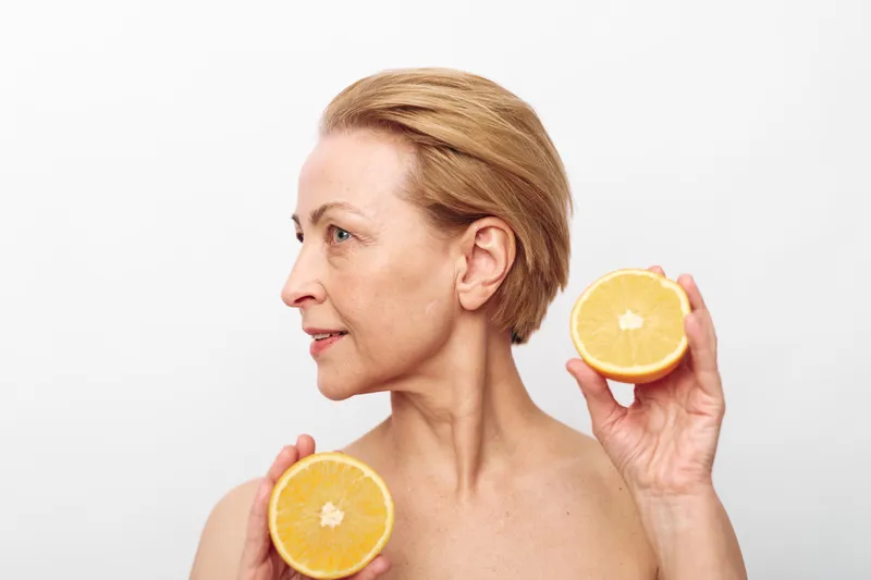 Mature woman with short hair holds two halves of fresh orange near her face on white background. Concept for healthy lifestyle, natural skincare, vitamin C, and wellness in beauty.