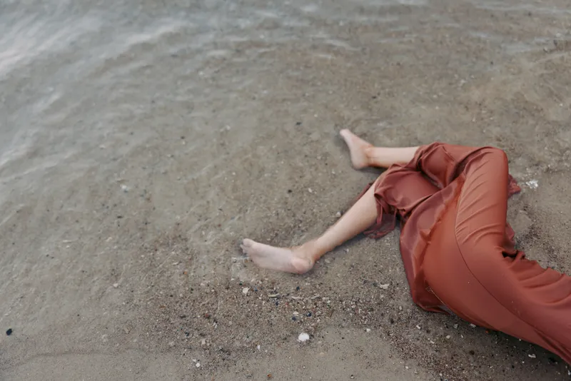 A figure lies on a sandy shoreline wearing a rust colored dress, bare feet in the wet sand as gentle waves touch the shore, creating a calm, contemplative coastal moment.