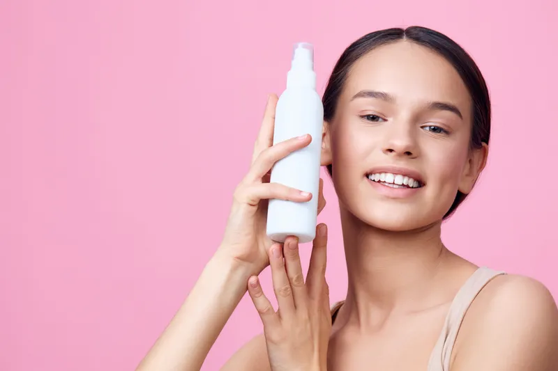 Young woman with a natural smile holding a skincare spray on a pink background, promoting healthy skincare and beauty routines