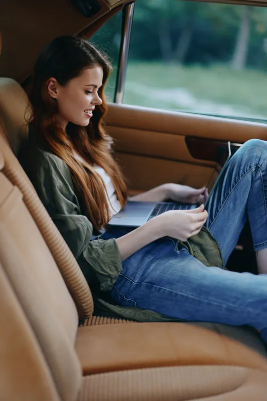 Relaxed young woman reading in a car, dressed casually in denim and a green jacket, exuding calmness and contentment with a natural outdoor background.