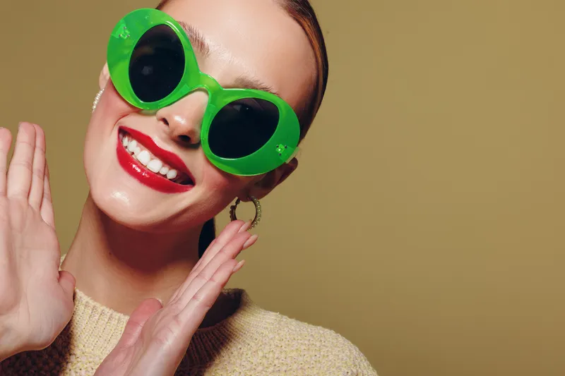 Smiling woman with bright red lipstick wears large green sunglasses and beige sweater, posing happily against plain background. Concept of fashion, summer style, and joyful mood.