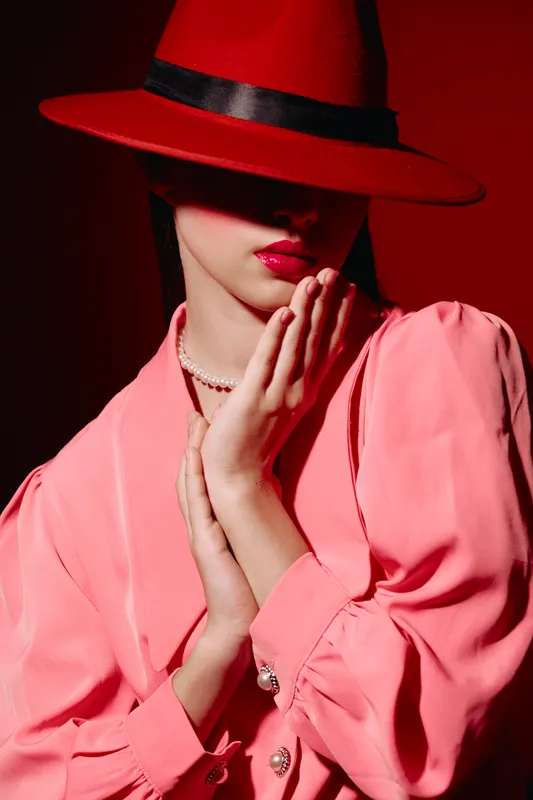 Fashionable woman in a red hat and pink blouse poses with hands near her face, lips painted red. Bold beauty and high-fashion portrait on a red background.