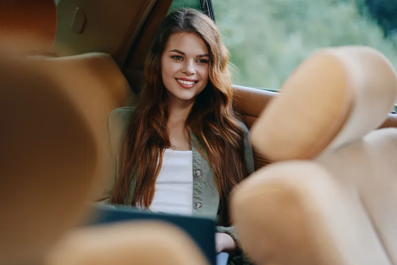 Young woman smiling in a car with a relaxed expression, dressed casually in a white top and green jacket, natural background, conveying happiness and comfort.