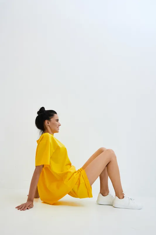 Young woman wearing bright yellow dress and white sneakers sitting on white floor in minimalistic studio, relaxed pose with casual style and soft lighting.