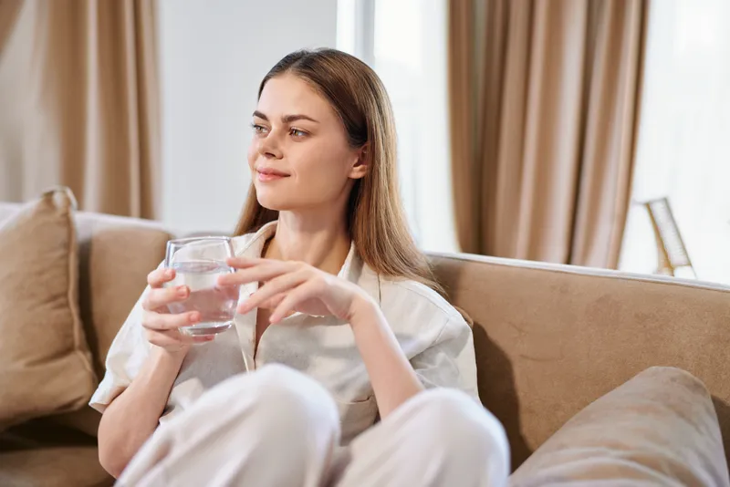 Woman sitting on sofa holding a glass of water at home, relaxed portrait in a cozy living room with natural light. Casual outfit, soft cushions and thoughtful calm expression for wellness.