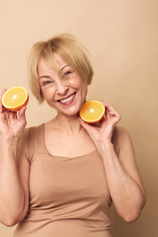 Happy middle aged woman with short blonde hair holds two orange halves near face smiling brightly on beige background. Concept of healthy lifestyle, vitamin c, wellness and natural beauty.