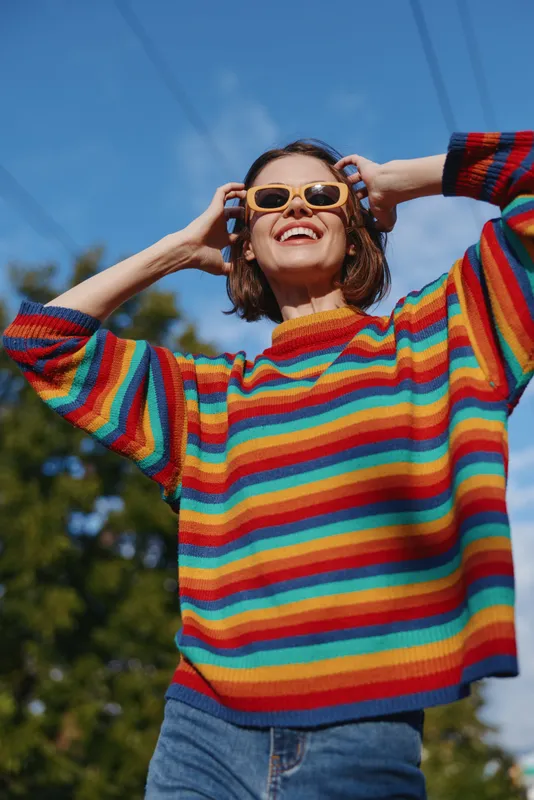 Woman in a colorful striped sweater and sunglasses smiling outdoors. Young woman in casual outfit adjusting glasses, short bob hair, joyful expression against blue sky for lifestyle fashion.